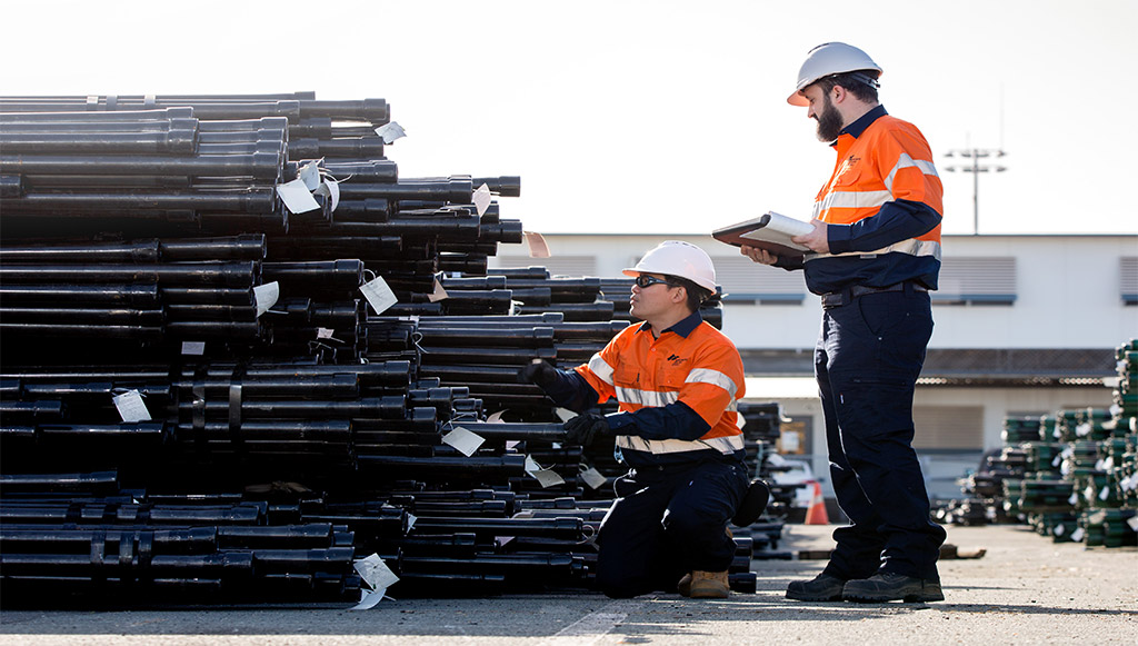 Two workers in safety gear assess pipes at a storage yard.